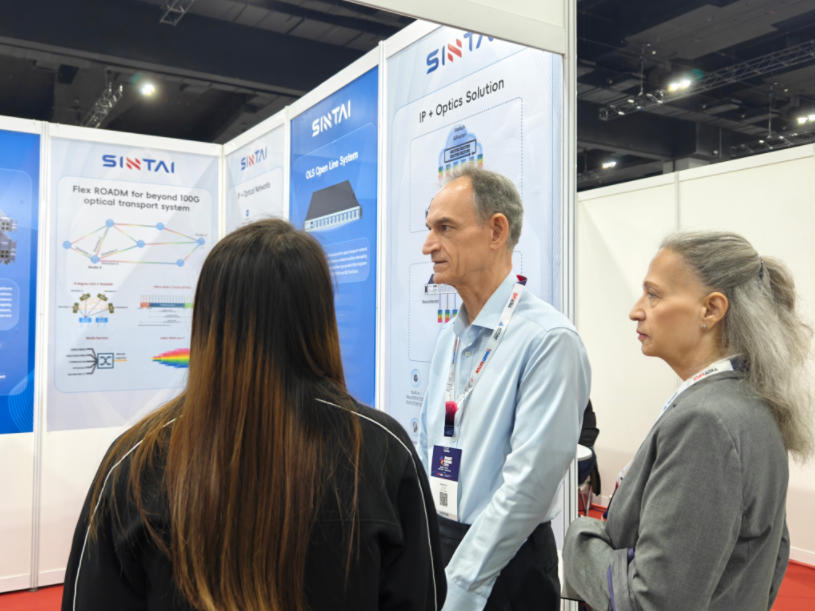 Two event attendees listening as a Sintai staff member speaks to them in front of the company's booth.